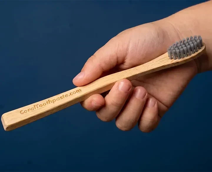 Hand holding a Coral bamboo toothbrush with soft gray bristles against a dark blue background.