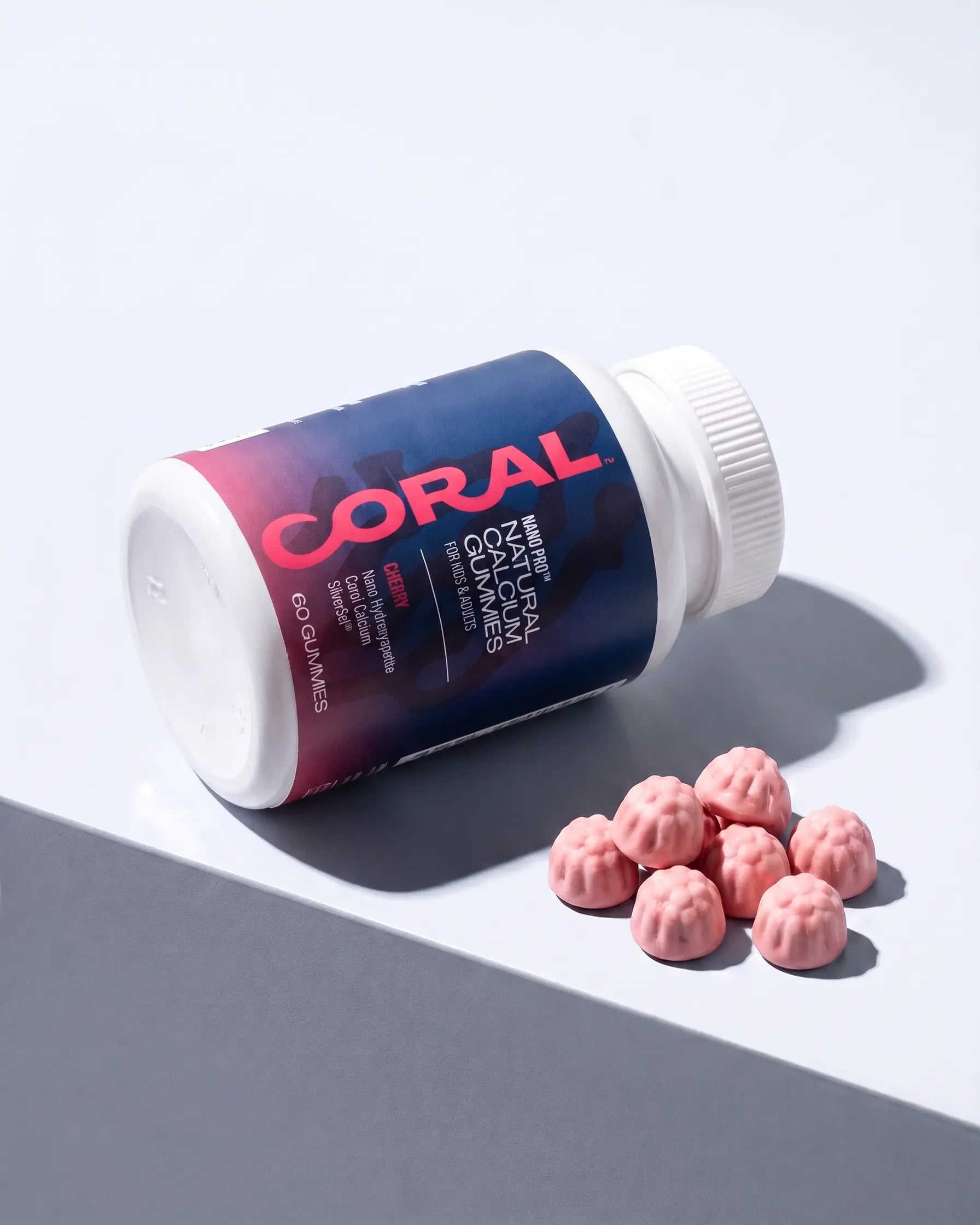 A white plastic bottle of Coral Nano Pro calcium gummies on its side next to several pink, textured gummies on a clean, light gray surface with a strong diagonal shadow.