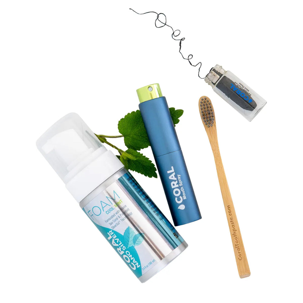 Flat lay of Coral natural oral care essentials including Coral Nano Silver Foaming Toothpaste, Coral Mouth Spray, bamboo toothbrush, and bamboo charcoal dental floss with mint leaves, showcasing eco-friendly and fluoride-free dental care products.