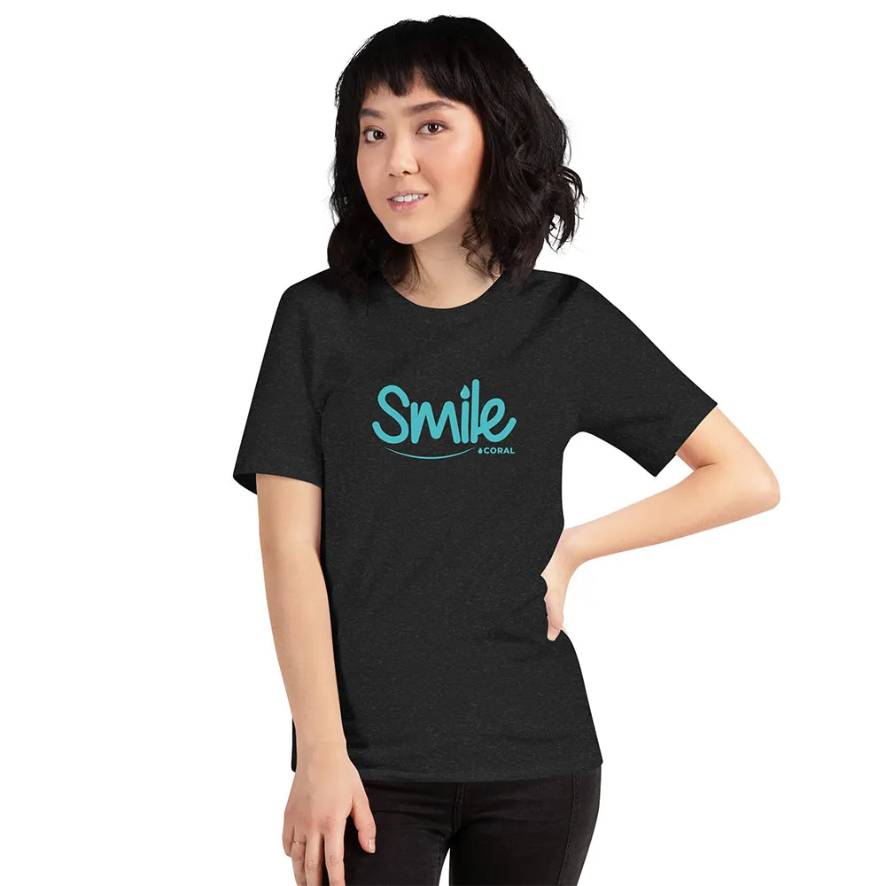 Woman wearing a black t-shirt with blue “Smile by Coral” logo text, standing against white background. Casual eco-friendly apparel promoting Coral oral care brand and confident, healthy smiles.